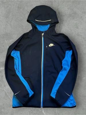 Nike blå och mörk blå  vindjacka med huva - Nike vindjacka i mörk blå och blått med huva och hel dragkedja framtill. Jackan har gul Nike-logga på bröstet, blå insida och detaljer i gult vid dragkedjan och huvan. Materialet är polyester och fleece, perfekt för träning utomhus. Reflekterande prickar på ärmarna.