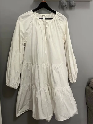 Vit volangklänning från H&M - Säljer en vit klänning från H&M i storlek S. Klänningen har lång ärm med resår vid ärmslut, volanger i flera lager och rund hals med knytband. Tillverkad i mjuk bomull som känns skön mot huden. 