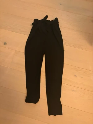 Svarta kostymbyxor från JDY med knyt - Snygga svarta kostymbyxor från JDY i storlek XS. Byxorna har hög midja och ett brett knytband i midjan som ger en cool touch. Modellen är rak och passar perfekt till en trendig look. Materialet känns mjukt och skönt mot huden.