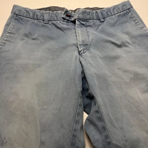 Ljusblå kostymbyxor från Oscar Jacobson - Snygga ljusblå kostymbyxor från Oscar Jacobson med klassisk rak passform. Byxorna har bälteshällor, knapp och dragkedja i midjan samt diskreta fickor. Materialet är mjukt och känns som bomull, perfekt för en stilren look.