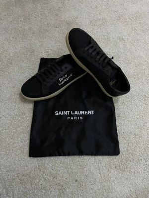 Saint Laurent Sneakers - Säljer ett par ÄKTA svarta sneakers från Saint Laurent med handskriven logga på sidan. Skorna har rund tå, platt sula och är tillverkade i canvas med svarta snören. Klassisk och stilren design med diskreta detaljer. Storlek 41 sitter som 41. Skick 10/10. Endast dustbagen och själva skorna medföljer.