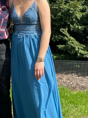 Blå långklänning med spetsdetaljer - Säljer en elegant blå långklänning med spets och broderade detaljer upptill. Klänningen har v-ringad urringning och är ärmlös med tunna axelband. Kjolen är i ett lätt och flödande material som ger en snygg siluett. Perfekt för bal 
