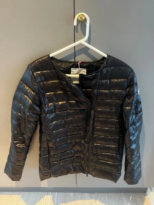 Svart pufferjacka från Moncler - Säljer en svart pufferjacka från Moncler med klassisk logga på ärmen. Jackan har quiltad design, rund hals och dragkedja framtill. Materialet är glansigt och känns lätt, perfekt för kyliga dagar. Som ni kan se på sista bilden så är själva dragkedjan utbytt för att den förra krånglade, men den medföljer och om du är kunnig kanske du kan sätta tillbaka den. Lm du har några frågor är det bara att fråga💕