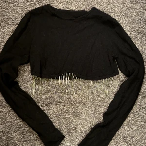 Svart croppad topp med pärlfransar - Säljer en svart croppad långärmad topp med glittrande pärlfransar längs nederkanten och vid ärmsluten. Toppen har en enkel rund halsringning och är tillverkad i mjukt tyg som känns bekvämt mot huden. Perfekt för dig som vill sticka ut med lite extra bling.