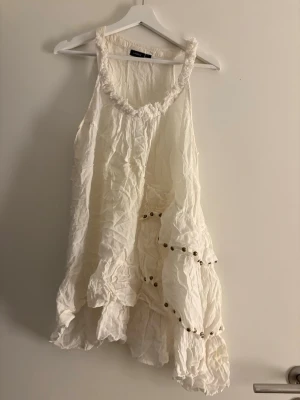 Vit volang linnetopp från Lindex - Säljer en vit linnetopp från Lindex i storlek L. Toppen har volanger och dekorativa metallnitar längs volangkanterna. Axelbandslös och med råa kanter runt halsringningen för en boho-vibe. Perfekt för varma dagar och sommarhäng.