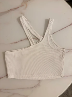 Vit ribbad oneshoulder topp - Säljer en vit ribbad topp med oneshoulder-design och dubbla smala axelband på ena sidan. Toppen är croppad och har en clean, modern look som passar perfekt till höga jeans eller kjol. Materialet är mjukt och stretchigt för skön passform.