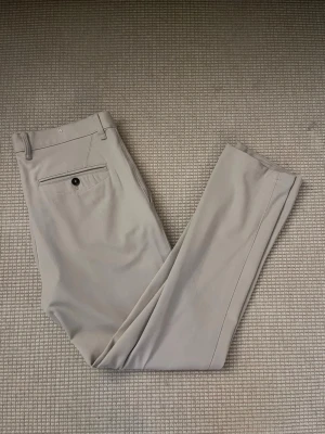 Beige kostymbyxor från Zara - Snygga beige kostymbyxor från Zara med raka ben och klassisk passform. Byxorna har bälteshällor, sidofickor och en bakficka med knapp. Tillverkade i ett mjukt bomullsmaterial som känns skönt mot huden.