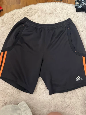 Svarta Adidas shorts med orange ränder - Säljer ett par svarta Adidas shorts med klassiska orangea ränder på sidorna och Adidas-logga framtill. Shortsen har elastisk midja och två fickor. Perfekta för träning eller chill dagar. Materialet är lätt och andas, troligtvis polyester.