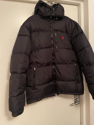 Svart pufferjacka från Polo Ralph Lauren - Svart pufferjacka från Polo Ralph Lauren med röd broderad logga på bröstet. Jackan har huva, flera dragkedjeförsedda fickor och justerbara muddar med kardborre. Materialet är polyester och jackan har en loose passform, perfekt för kalla dagar.Jackan är endast använd ett fåtal gånger och i mycket fint skick.