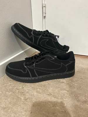 Nike Air Jordan 1 Low svart sneakers - Svarta Nike Air Jordan 1 Low sneakers med vit kontrastsöm och broderade detaljer på hälen. Skorna har rund tå, platt sula och klassisk snörning. Materialet är skinn och syntet, och yttersulan har tydligt mönster för bra grepp. Perfekt för dig som gillar stilrena och ikoniska sneakers.