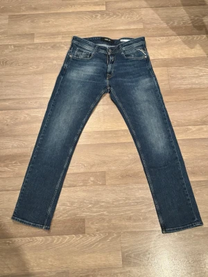 Replay Rocco blå jeans - Snygga Replay Rocco jeans i klassisk blå tvätt med diskreta slitningar. Modellen har raka ben, fem fickor och detaljerade sömmar på bakfickorna. Materialet är slitstark jeansbomull och passformen är normal. Perfekt för dig som gillar stilrena och tidlösa jeans.