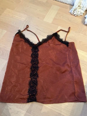 Brun spetsdetaljerad linnetopp Chiara Forthi Milano - Säljer en brun/orange linnetopp från Chiara Forthi Milano med tunna axelband och snygga svarta spetsdetaljer längs urringningen och mitt fram. Toppen har en mjuk, lite glansig yta och är perfekt för dig som gillar feminina detaljer.