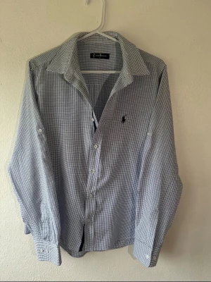 Rutig skjorta från Ralph Lauren - Snygg rutig skjorta från Ralph Lauren i blått och vitt. Klassisk krage, långärmad och med knappar framtill. Skjortan har det ikoniska Polo-logotypen broderad på bröstet. Perfekt för dig som gillar stilrena och tidlösa plagg.