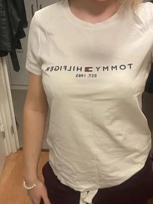 Vit t-shirt från Tommy Hilfiger - Säljer en vit t-shirt från Tommy Hilfiger med klassisk logga och texten 'EST. 1985' på bröstet. T-shirten har rund hals och korta ärmar, samt en liten logga på ärmen. Materialet är mjuk bomull och passformen är normal.