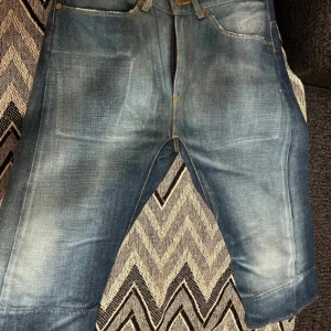 Levi's 501 blå jeans med rak passform - Säljer ett par klassiska Levi's 501 jeans i blå tvätt med snygga slitningar och kontrastsömmar. Modellen har rak passform, fem fickor och knappgylf. Jeansen är tillverkade i robust bomullsdenim och har den ikoniska röda Levi's-taggen på bakfickan.