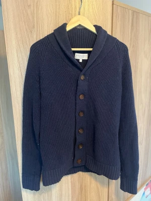 Mörkblå cardigan/kofta från William Hunt - Säljer en mörkblå, grovstickad kofta eller cardigan från William Hunt(savile row) i storlek Medium. Perfekt för lager-på-lager under kyliga dagar. Använd varsamt och förutom lite nopprig inga slitagetecken.