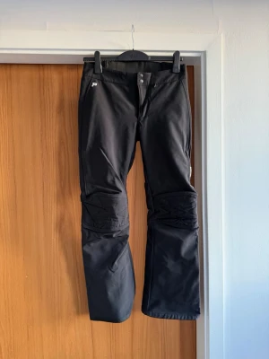 Peak performance Svarta Sofshell  - Säljer ett par svarta täckbyxor från Peak Performance. Byxorna har quiltade detaljer vid knäna, dragkedjor vid bensluten och två knappar i midjan. Materialet är slitstarkt och passar perfekt för vinterns aktiviteter.
