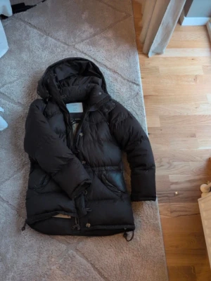 Svart dunjacka med huva - Säljer en svart dunjacka från Clair med stor, mysig huva och dragkedja framtill. Jackan har quiltad design, långa ärmar och praktiska fickor på sidorna. Perfekt för kalla dagar och har justerbar nederkant med snörning.