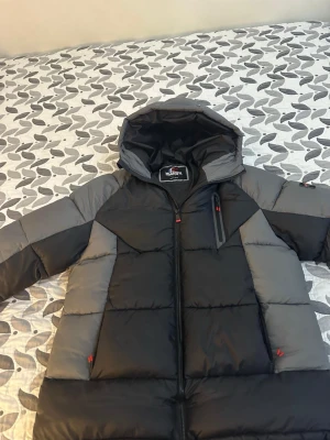 Svart och grå pufferjacka McKenzie - Säljer en fet pufferjacka från McKenzie i svart och grått med huva och coola röda detaljer på dragkedjorna. Jackan har snörning nedtill och en bröstficka med dragkedja. Perfekt för kalla dagar när du vill hålla stilen.