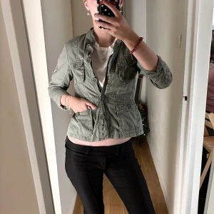 Grön utilityjacka med fickor - Snygg grön utilityjacka med flera stora fickor framtill och knappstängning. Jackan har en ståkrage och är tillverkad i bomull, vilket gör den både bekväm och lätt att bära. Perfekt för dig som gillar en avslappnad men cool stil. Barnstorlek men passar kanske en xxs/xs