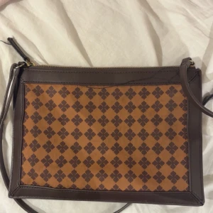 Brun axelväska från By Malene Birger - Snygg axelväska från By Malene Birger med brunt och beige rutigt mönster. Väskan har en praktisk dragkedja upptill och justerbar axelrem. Materialet är en mix av skinn och textil, med klassisk logodetalj framtill. Perfekt storlek för det viktigaste.