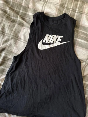 Svart linnetopp från Nike - Svart linnetopp från Nike med stort vitt logotryck på bröstet. Modellen är ärmlös och har rund halsringning. Tillverkad i mjuk bomull som känns skön mot huden. Perfekt för träning eller chill dagar.