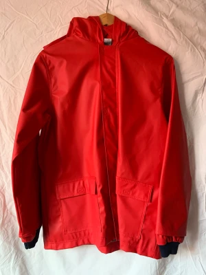 Röd regnjacka från Petit Bateau - Snygg röd regnjacka för barn från Petit Bateau med huva och två stora fickor framtill. Dragkedja och kardborreband. Insidan har ett mörkblått och vitt randigt foder i bomull som känns skönt mot kroppen. Jackan är gjord i syntetmaterial och har långa ärmar med mörkblå muddar. Aldrig använd så i perfekt skick. Nypris ca 1300 kr. 12 år/152 cm men liten fransk storlek!