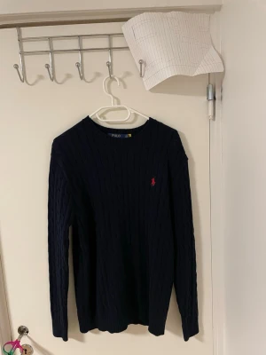 Mörkblå stickad tröja  Polo Ralph Lauren - Säljer en mörkblå stickad tröja från Polo Ralph Lauren med klassiskt kabelstickat mönster och rund hals. Tröjan har lång ärm och en liten röd broderad logga på bröstet. Perfekt för dig som gillar stilrena och tidlösa plagg.Liknande modell som orginala. 
