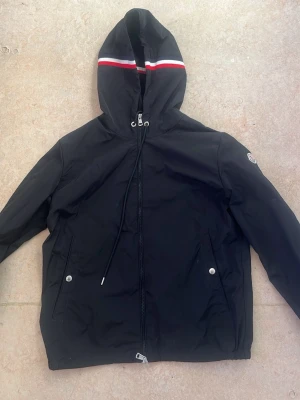 Svart vindjacka från Moncler - Svart vindjacka från Moncler med huva som har snygga ränder i rött, vitt och mörkblått. Jackan har dragkedja framtill, snörning vid huvan och två fickor med silverfärgade knappar. Perfekt för dig som gillar stilrena och sportiga plagg. NFC✅