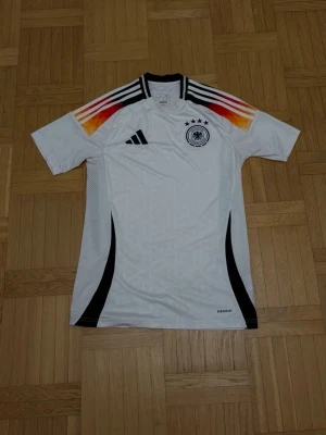 Vit Adidas Tyskland fotbollströja S - Säljer en officiell Tyskland fotbollströja från Adidas i storlek S. Tröjan är vit med svarta detaljer, har slim fit passform och coola ränder i rött, orange och svart på axlarna. Broderat Tysklands emblem med fyra stjärnor på bröstet och Aeroready-material för extra komfort.