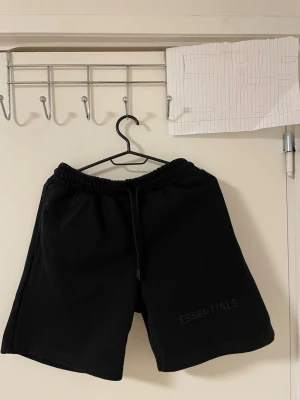 Svarta Essentials shorts   - Svarta shorts från Essentials med elastisk midja och snörning framtill. Mjukt material och enkel design med diskret logga på benet. Har en bakficka och avslappnad passform som är perfekt för chill dagar eller träning.