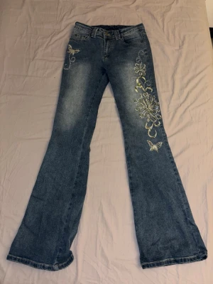 Blå bootcut jeans med broderi strlk 38w - Säljer ett par blå bootcut jeans med snygga broderade detaljer och paljetter på ena benet. Jeansen har blommönster och fjärilar i broderi, klassisk femficksdesign och bälteshällor. Materialet är jeans och passformen är loose med utsvängda ben. STRLK 38W kan mötas I stockholm