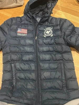 Svart pufferjacka från Vinson Polo Club - Snygg svart pufferjacka från Vinson Polo Club med broderade detaljer och logga på bröstet. Jackan har huva, dragkedja framtill och quiltad design. Perfekt för dig som gillar sportig stil och vill hålla dig varm under kalla dagar.