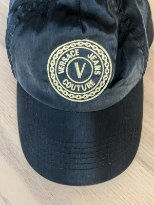 Svart Versace Jeans Couture keps - Snygg svart keps från Versace Jeans Couture med broderad logga i guld framtill. Kepsen har en klassisk form och är tillverkad i syntetmaterial som ger en lätt och bekväm känsla. Perfekt accessoar för att lyfta din streetwear-look.
