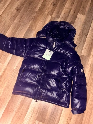 Mörkblå Moncler pufferjacka M - Säljer en mörkblå pufferjacka från Moncler i storlek M. Jackan har glansig yta, huva och dragkedja framtill samt två sidofickor med dragkedja. Insidan har ett coolt serietryck och klassisk Moncler-logga på ärmen. Perfekt för kalla dagar.