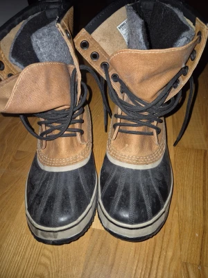 Sorel Caribou kängor i skinn och gummi - Bruna och svarta Sorel Caribou kängor med snörning, tillverkade i skinn och gummi. Fodrade med grå filt för extra värme och har en rejäl platt sula. Perfekta för kalla dagar och tuffa väderförhållanden.