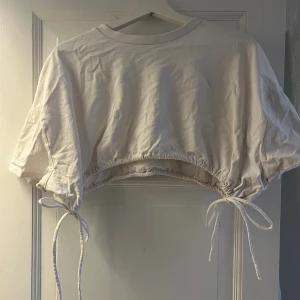 Vit croppad topp med knyt - Trendig vit croppad topp med korta ärmar och snygga knytband i sidorna. Toppen har en avslappnad passform och är gjord i mjuk bomull, perfekt för en chill och stilren look.