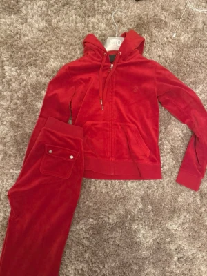 Röd velour loungewear-set Juicy Couture - Säljer ett ikoniskt rött loungewear-set från Juicy Couture i mjuk velour. Setet består av en zip-hoodie med dragsko och fickor samt matchande byxor med resårmidja, dragsko och fickor med knapp. Perfekt för chill dagar hemma eller när du vill vara extra comfy. Nypris 2300. som nytt