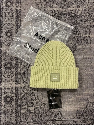 Acne Studios beanie  - Säljer en ribbstickad ljusgrön mössa från Acne Studios med den ikoniska smiley-loggan framtill. Mössan har uppvikt kant och är tillverkad i mjukt material som håller dig varm och snygg under kalla dagar.