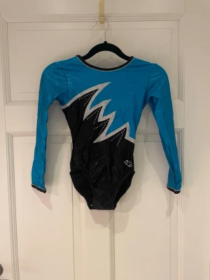 Blå och svart gymnastikdräkt - Säljer min gamla tävlingsdräkt till AG gymnastik då den blivit för liten. Dräkten har ett coolt blixtmönster i silver med små glittriga stenar på framsidan följt av samma blixtnömster på baksidan. Perfekt för träning eller tävling inom gymnastik. Storlek 140-152. Nypris är 2199 kr.