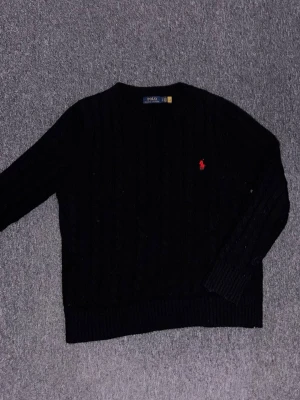 Svart kabelstickad tröja Polo Ralph Lauren - Kabelstickad svart tröja från Polo Ralph Lauren med rund halsringning och den klassiska röda logotypen broderad på bröstet. Tröjan har ribbade muddar vid ärmslut och nederkant, perfekt för en stilren och tidlös look.