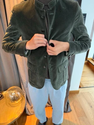 Mörkgrön manchesterkavaj med ståkrage - Snygg mörkgrön kavaj i manchester med ståkrage och knappar framtill. Två stora fickor sopm stängs med knapp och en minimalistisk design utan slag. Perfekt för dig som vill ha en unik twist på klassisk kavajstil.