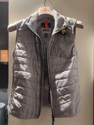 Ljusgrå dunväst från Parajumpers - Snygg ljusgrå dunväst från Parajumpers med quiltad design och hög krage. Västen har dragkedja framtill, meshdetaljer vid axlarna och en gul detalj vid halsen. Perfekt för lager-på-lager och håller dig varm under kyliga dagar.