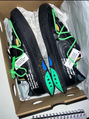 Nike Blazer Low x Off-White svart/grön - Nike Blazer Low x Off-White sneakers i svart med gröna och blå detaljer samt gröna snören. Skorna har unika Off-White-texter på sidorna och en chunky svart sula med färgstänk. Materialet är skinn och syntet, och designen är modern och edgy.