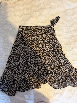 Dam Leopardmönstrad kjol - Säljer en lång leopardmönster i beige och svart. Kjolen har volangkant nertill och knytband i midjan för extra detalj i storlek 134/140 men passar mig som har XS/XXS. Perfekt för dig som vill sticka ut med ett djurmönster och gillar trendiga detaljer.