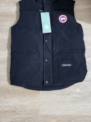 Svart dunväst  - Stilren svart dunväst med klassisk logga på bröstet och två stora fickor framtill. Västen har tryckknappar och en rak passform, perfekt för lager på lager under kyliga dagar. Materialet är slitstarkt och håller värmen riktigt bra.