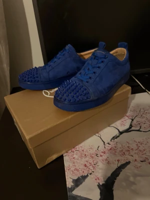 Blå mocka sneakers med nitar från Christian loubetins  - Snygga blå sneakers i mocka med coola nitar på tån och snörning. Skorna har en rund tå och platt sula, vilket ger en modern och edgy look. Perfekta för dig som vill sticka ut med din stil. Bara att skriva om du har några frågor skorna är i 43/42 