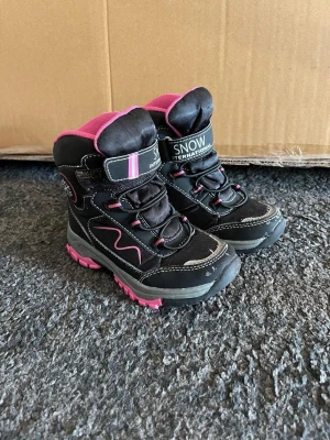 Svarta vinterstövlar med rosa detaljer - Säljer ett par svarta vinterstövlar med rosa detaljer och grov sula. Stövlarna har kardborreband upptill, snörning framtill och är fodrade för extra värme. Märket är Snow International. Perfekta för kalla dagar!
