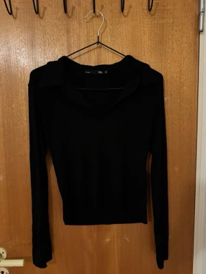 Svart långärmad tröja med krage - Stilren svart långärmad tröja med klassisk krage och v-ringning. Tröjan har en smal passform och är tillverkad i ett mjukt, stretchigt material som passar perfekt till både jeans och kjol.