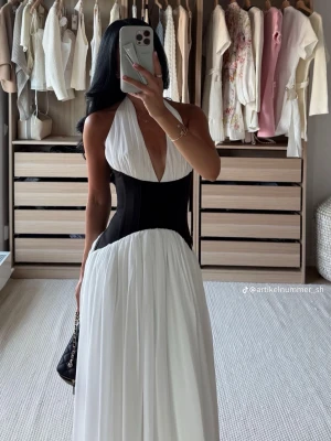 Vit långklänning med svart midja - Elegant vit långklänning med djup v-ringning och markerad svart midja som ger en snygg siluett. Klänningen har halterneck och ett flytande, plisserat tyg som faller mjukt. Perfekt för dig som vill sticka ut med en stilren look.